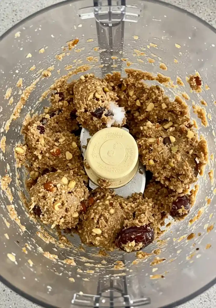 Food processor bowl with dates, coconut oil and vanilla being added to dry ingredients mixture for chocolate energy balls