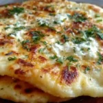 Stack of golden-brown cottage cheese flatbreads on wooden cutting board with herbs and olive oil