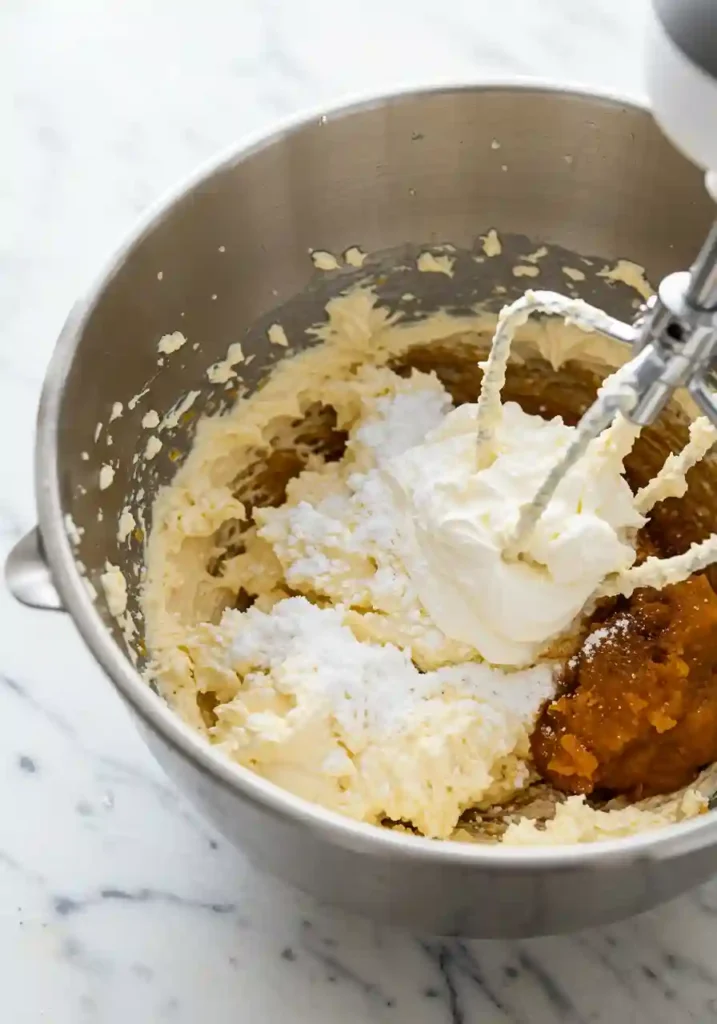 Electric mixer beating orange pumpkin cheesecake filling in large mixing bowl showing smooth creamy texture