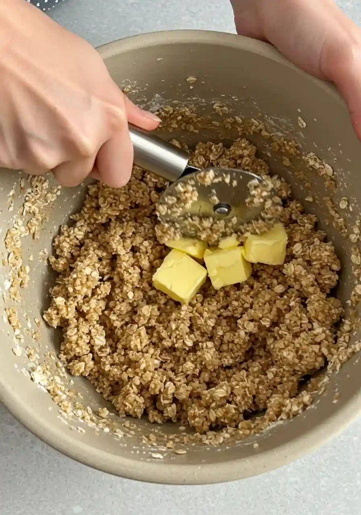 Hands cutting cold butter into oat flour mixture with pastry cutter showing proper crumb texture for Apple Crisp Bars