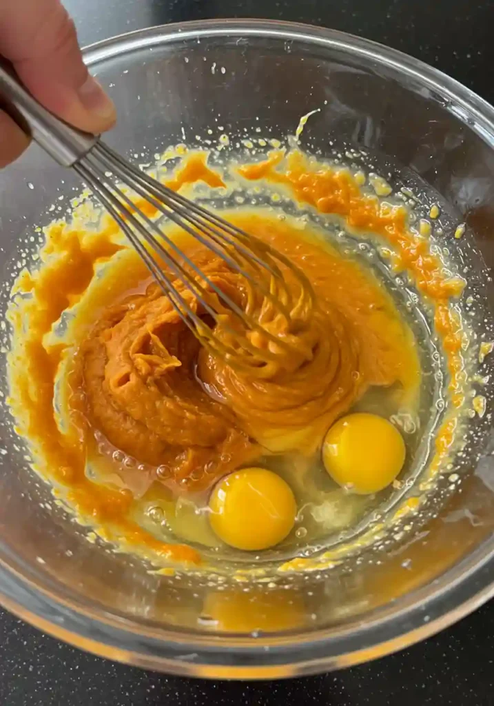 Hands whisking pumpkin puree with eggs and oil in glass bowl for moist pumpkin bread batter