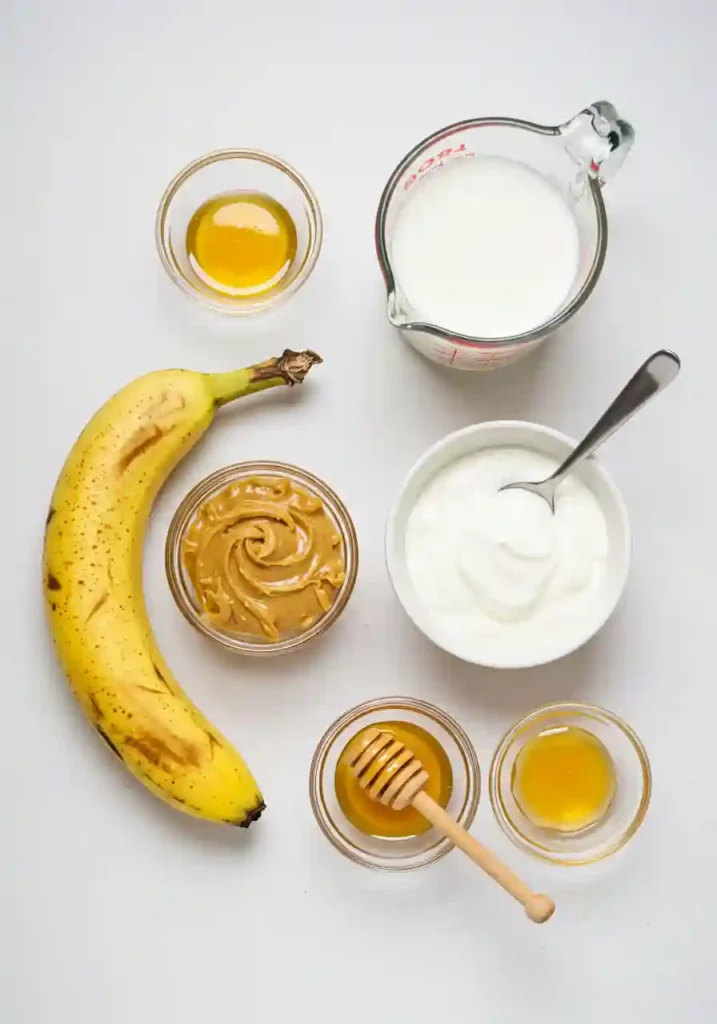All ingredients for banana peanut butter smoothie including banana, Greek yogurt, peanut butter, milk, and honey arranged on white table
