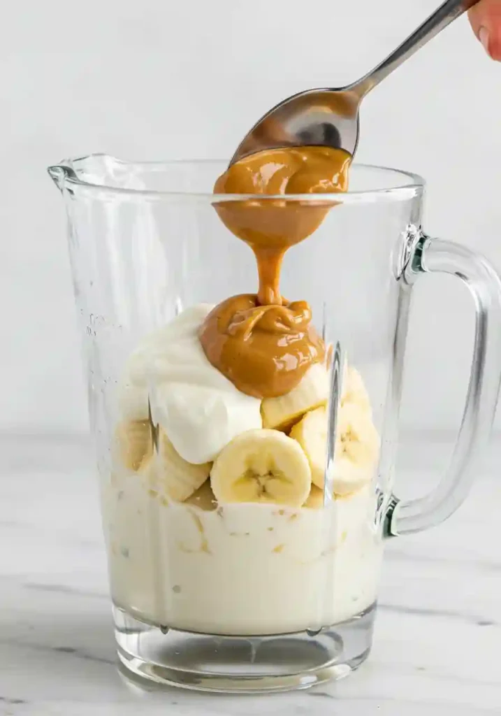 Layering milk, yogurt, banana, and peanut butter into blender jar for smoothie preparation