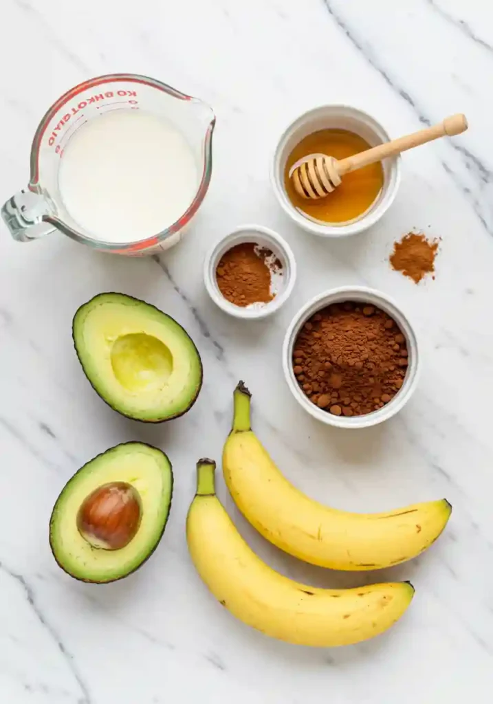 Flat lay of ingredients for chocolate avocado banana smoothie including ripe avocado, frozen bananas, cocoa powder, milk, and cinnamon on white marble