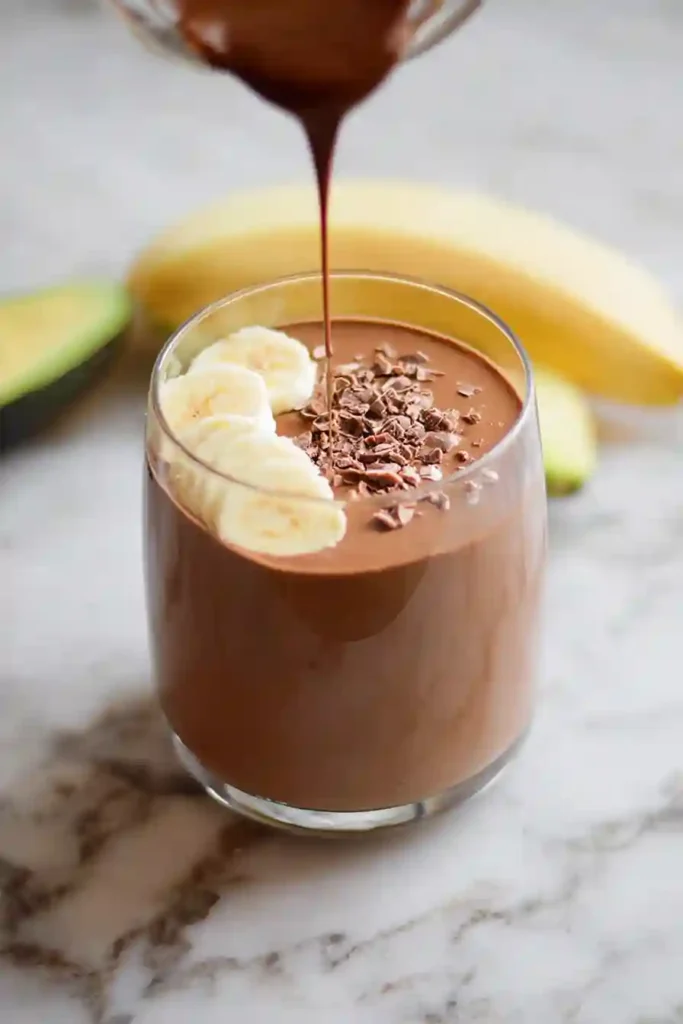 Thick chocolate avocado banana smoothie being poured from blender into clear glass showing creamy velvety texture