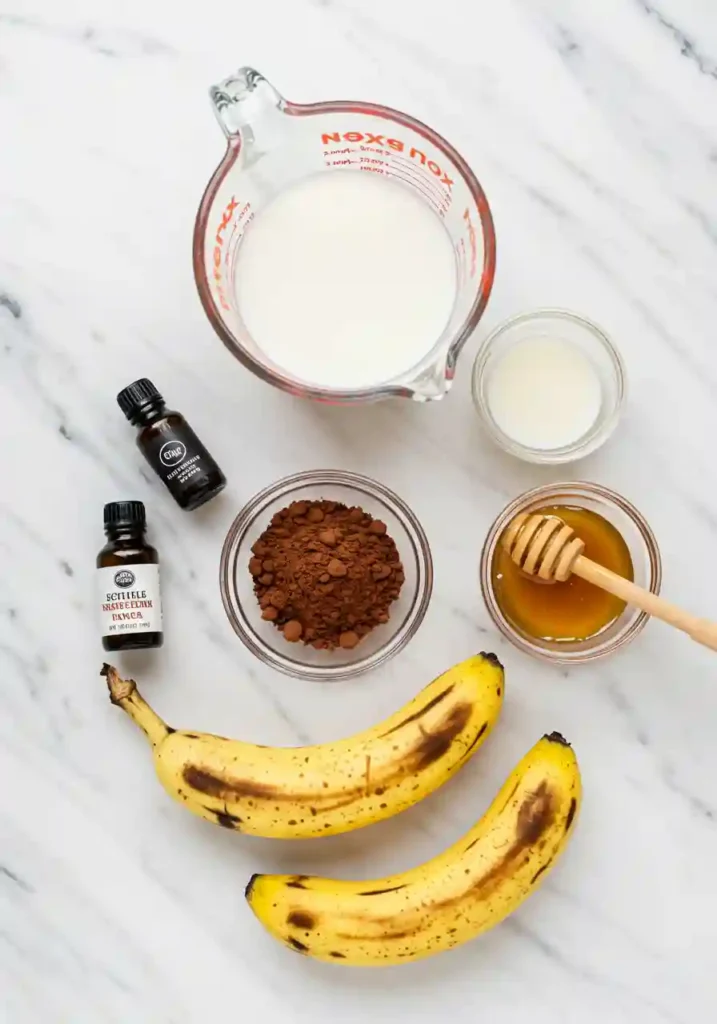 Chocolate banana smoothie ingredients including ripe bananas, cocoa powder, milk, vanilla extract, and honey arranged on counter