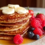 Stack of fluffy cottage cheese oatmeal pancakes drizzled with maple syrup, topped with butter and surrounded by fresh blueberries and raspberries